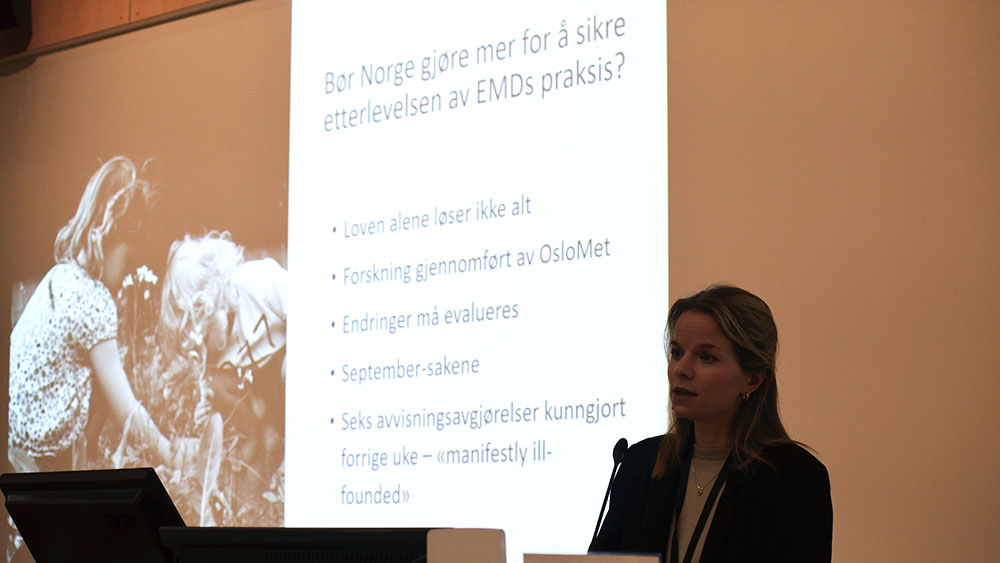 Bildet viser en kvinne som snakker. Kvinnen står ved et podium. I bakgrunnen er det en powerpoint-presentasjon med teksten "Bør Norge gjøre mer for å sikre etterlevelsen av EMDs praksis?".