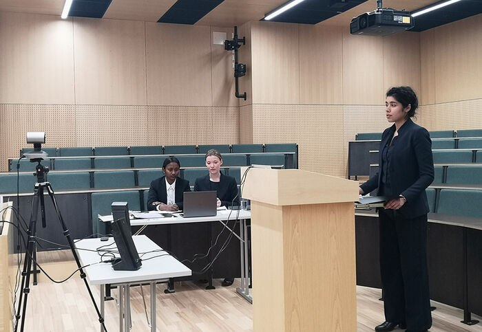 En student står foran et kamera og prosederer, mens to andre studenter sitter i bakgrunnen og følger med