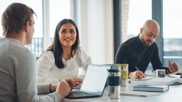 Tre studenter sitter rundt bord med PCer og diskuterer. 