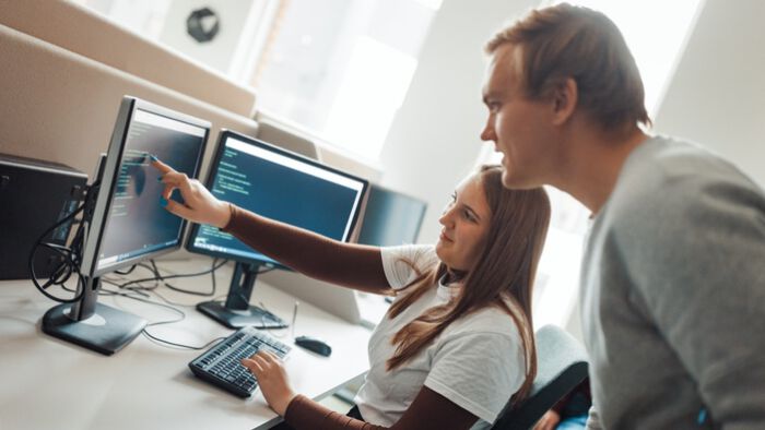 En student sitter foran en datamaskin og får hjelp til programmering av en veileder som står bak.