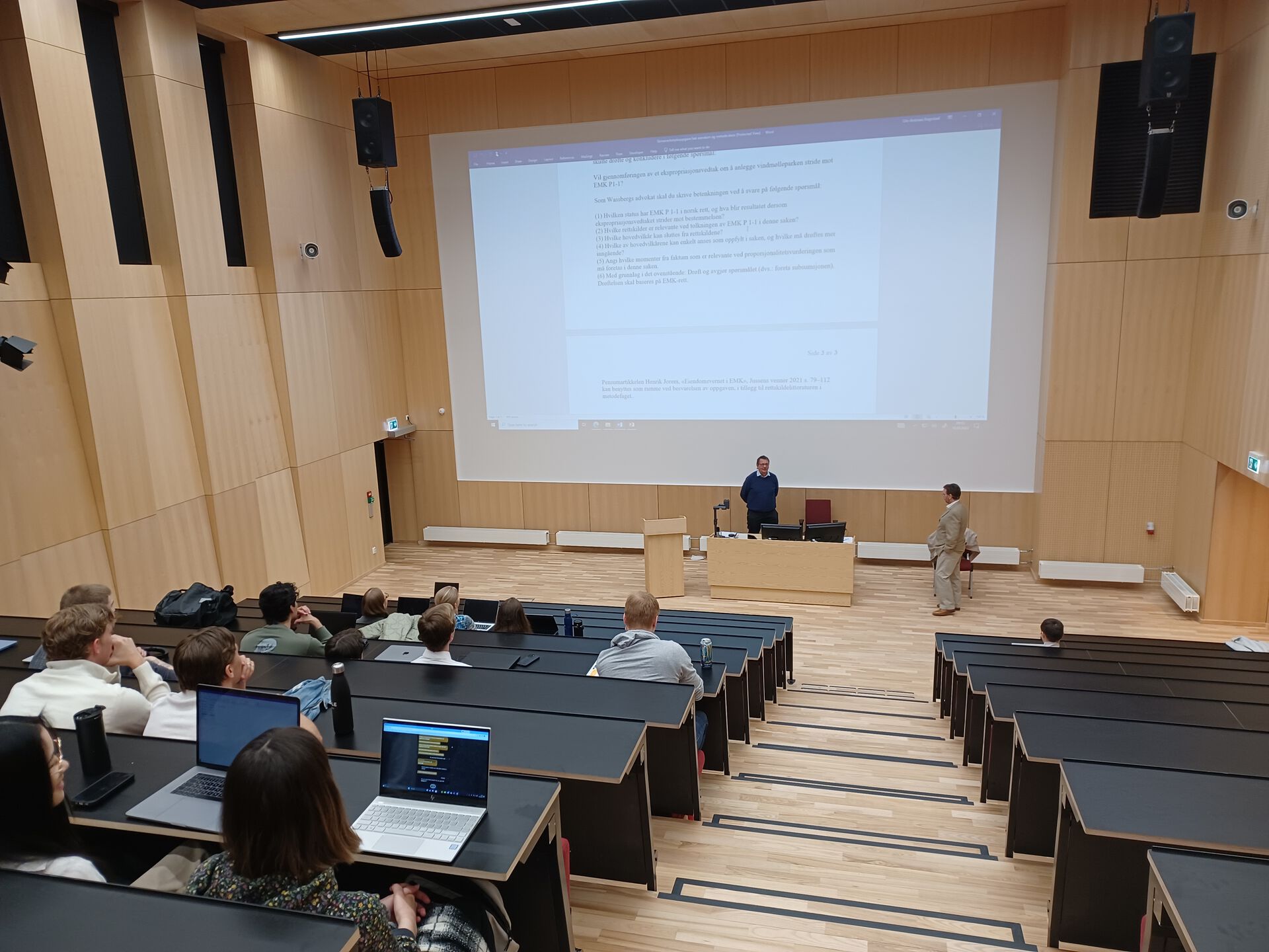 To menn står foran en storskjerm, man kan se baksiden av studenter som sitter i forelesningssalen.
