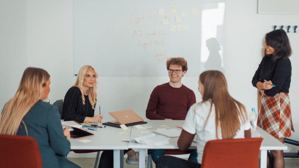 Bildet viser 4 studenter som sitter rundt et bord og 1 læringsassistent som står ved siden av og veileder. 