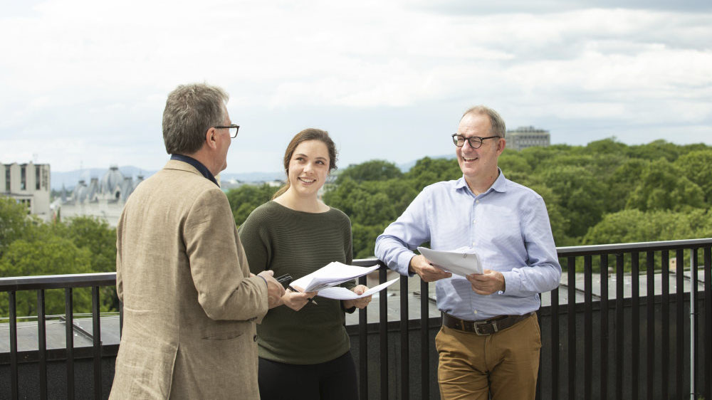 Image of two men and a woman on a rooftop, holding papers. One of the men has his back turned to the camera, seemingly talking to the other two.