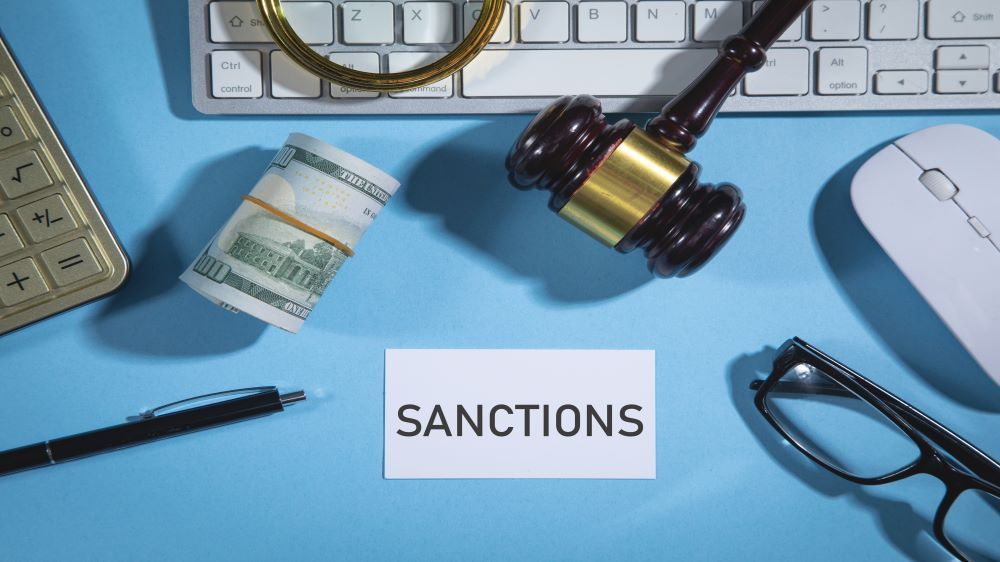 Illustration photo of a desktop. On top of the desktop there is a keyboard, computer mouse, glasses, justice hammer, glasses, American Dollars, a pen and a sticker saying "Sanctions". 