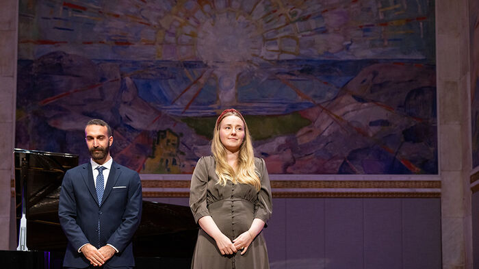 Ung dame står foran på scene med et Munch-maleri i bakgrunnen. Mann i dress til venstre.