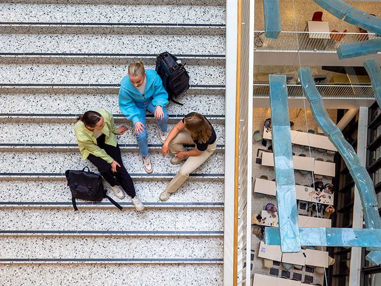 Bildet viser tre studenter som sitter i trappen og snakker sett ovenfra. På høyre side ser du ned til første etasje i Domus Juridica.