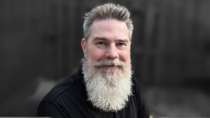 Photograph of Heith Copes against a blurry, dark background. Copes is a middle-aged white man with grey hair, a full white beard and mustache