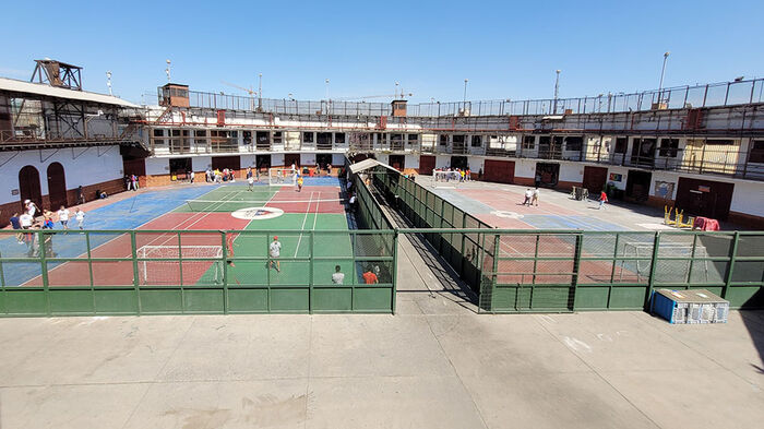 Outdoor area inside a prison with marked tennis courts on the ground and football goals. Several prisoners in various activities.