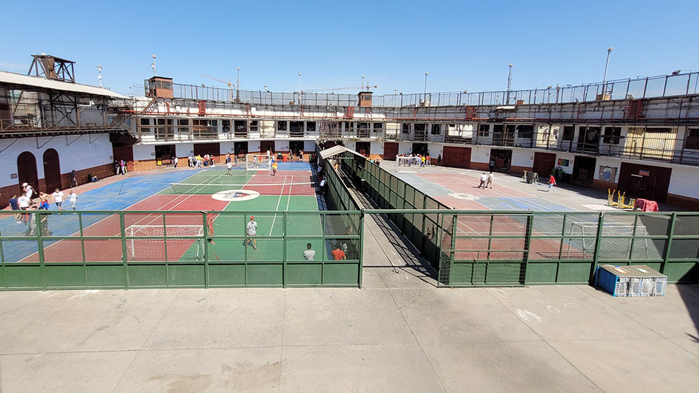 Uteområde inne i et fengsel med oppmerkede tennisbaner på bakken og fotballmål. Flere fanger i ulike aktiviteter. 