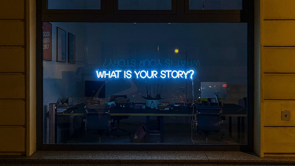 Et fotografi tatt fra gaten på kvelden og inn mot et mørkt og lite kontorlanskap. På vinduet henger det lysrør formet med i teksten "Hva er din fortelling?".
