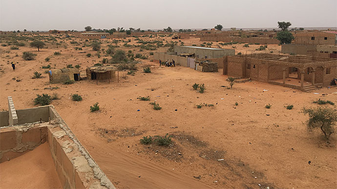 Afriskansk steppelandskap med noen trær, busker og mye sand. Bildet viser noen enkle huskonstruksjoner noen mennesker langt unna. 