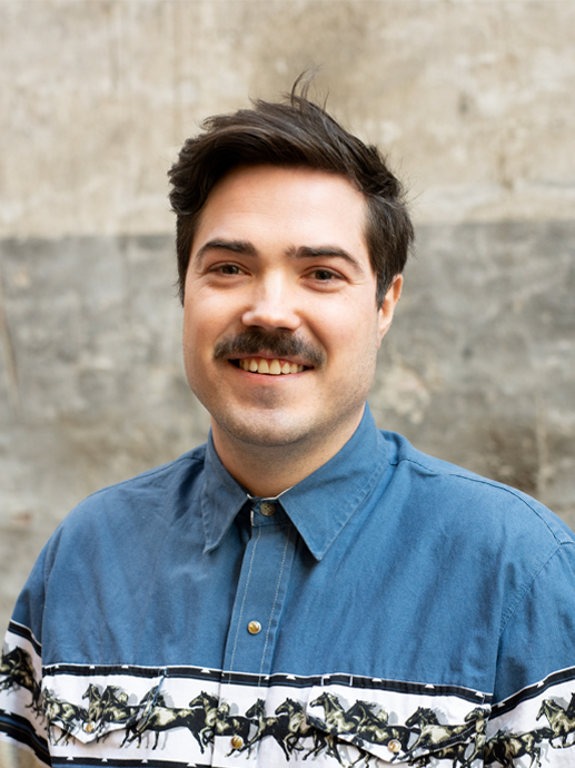 Photo of Vegard Haukland, a young white man with dark hair and a mustache, wearing a blue denum shirt with a pattern across it. 
