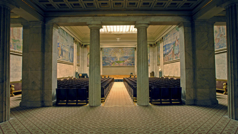 Bilde av søyler som holder taket oppe ved inngangen til aulaen. Veggmalerier av Munch i bakgrunnen.  Arkitektur, Interiørdesign.