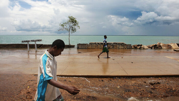 Two Torres Strait islanders walking down a flooded road, near the ocean.