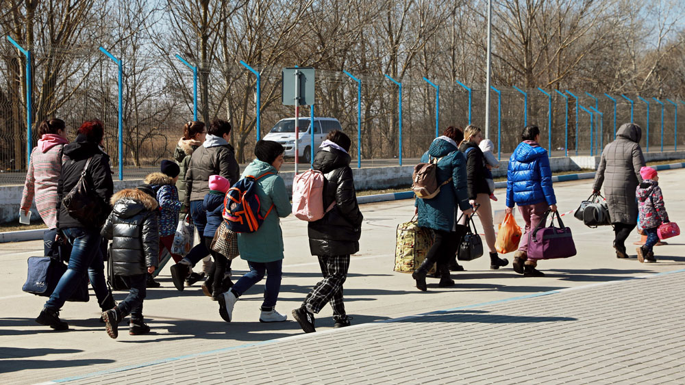 En gruppe kvinner og barn gående langs et grensegjerde, bærende på flere bager og nett