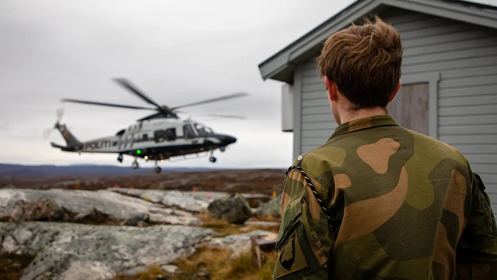 Et politihelikopter går inn for landing ved en grå hytte i et åpent landskap. Med ryggen til står en uniformskledd mann med emblemet til garnisonen i Sør-varanger på ermet.