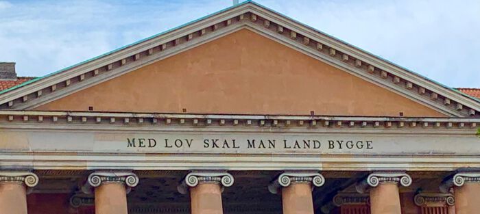 Bildet av storslått fasade med søyler og teksten «Med lov skal man land bygge» skrevet over søylene.