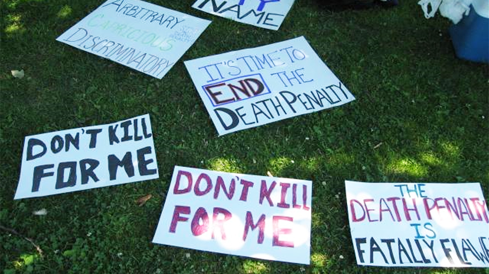 Several protest signs laying on the grass. They read: Don't kill for me, it's time to end the death penalty, the death penalty is fatally flawed.
