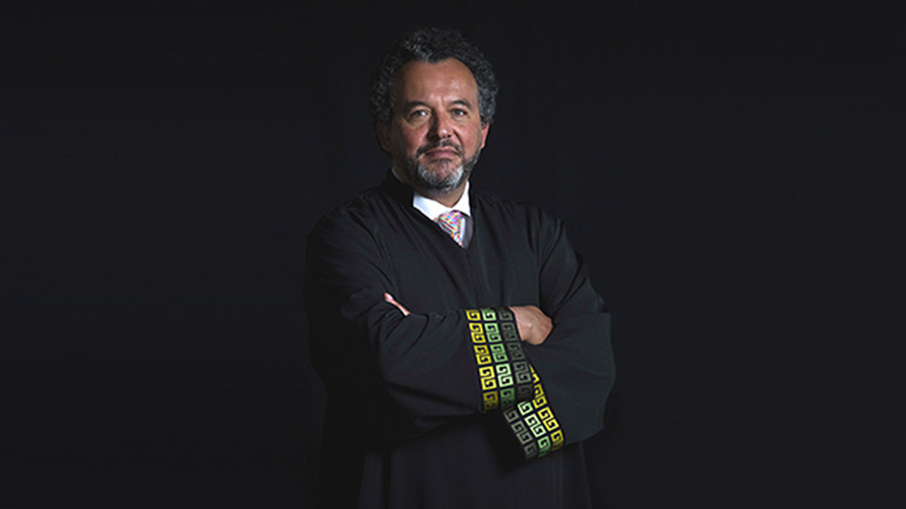 Man standing with his arms crossed in front of a black screen, looking serious. He is wearing a tie, black judge robe and has a beard.