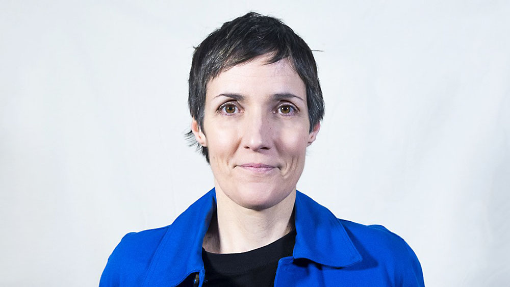 This image shows a woman with short hair and a blue jacket, standing in front of a white background. 