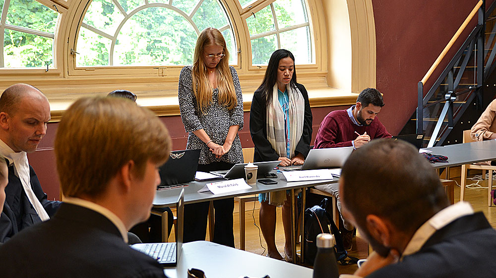 Two course participants facing a panel of judges, who have their backs to the camera. A large window in the background.