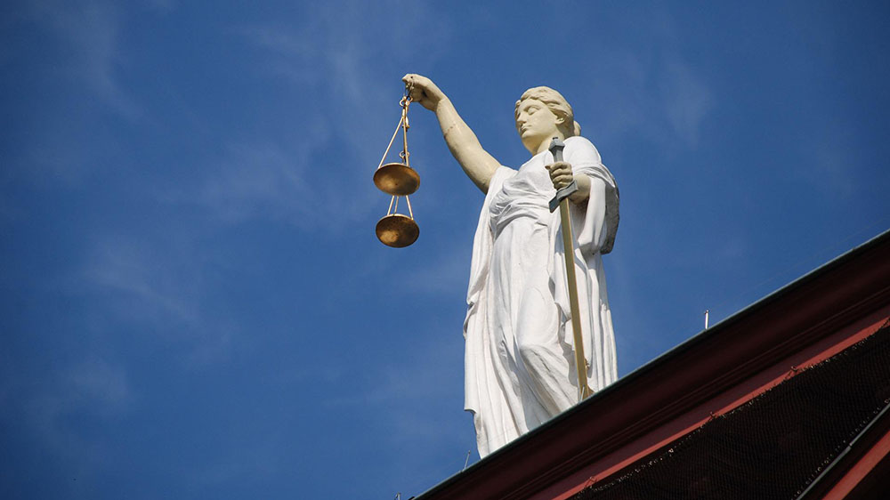 Image of a white "Lady Justice" statue holding scales in one hand and a sword in another, against a blue sky background