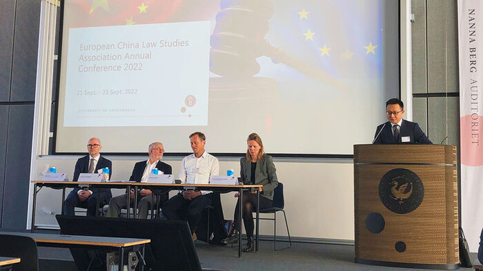 Image of a panel consisting of four people sitting behind a table in front of a PowerPoint presentation, as well as a speaker at a podium