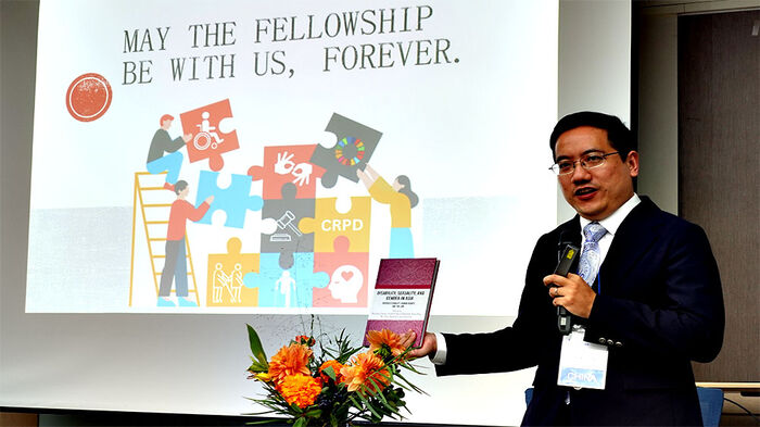 Image contains a professor presenting a book, talking in microphone with the book in his hand