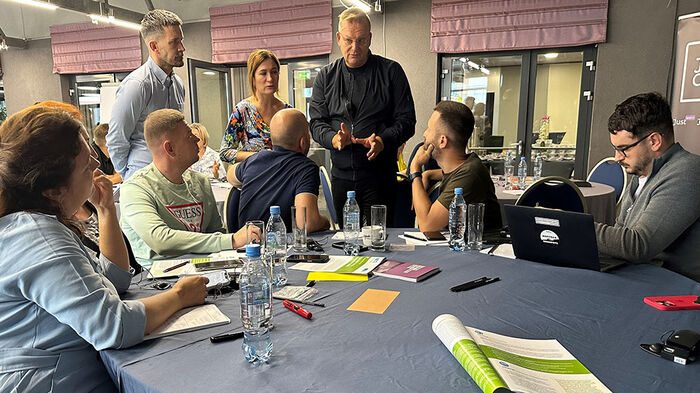 People sitting around a round table listening to one person who is standing. 