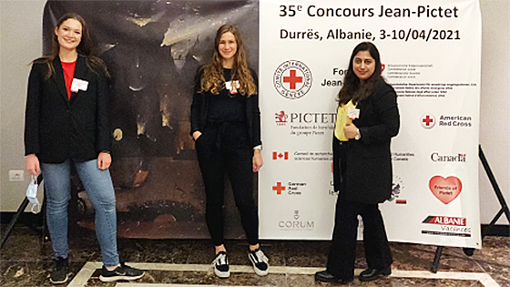Three participants, dressed in formalwear, in front of a Jean Pictet Moot Court Competition banner with various sponsor logos on it.