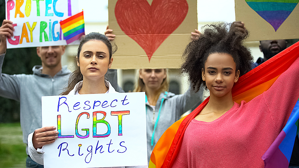 Pride-parade. To personer står foran en gruppe som holder regnbueflagg og plakater. Personen til venstre holder en plakat hvor det står "respect LGBT rights". Personen til høyre holder et stort regnbueflagg.